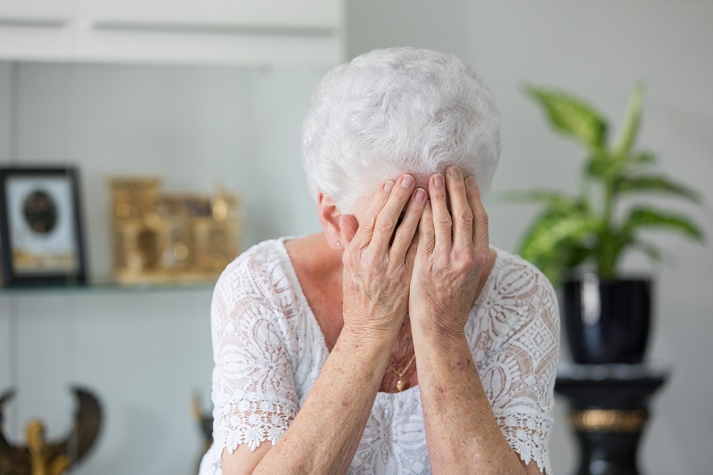 Een mevrouw met grijs haar. Ze zit en heeft haar handen voor haar gezicht. 